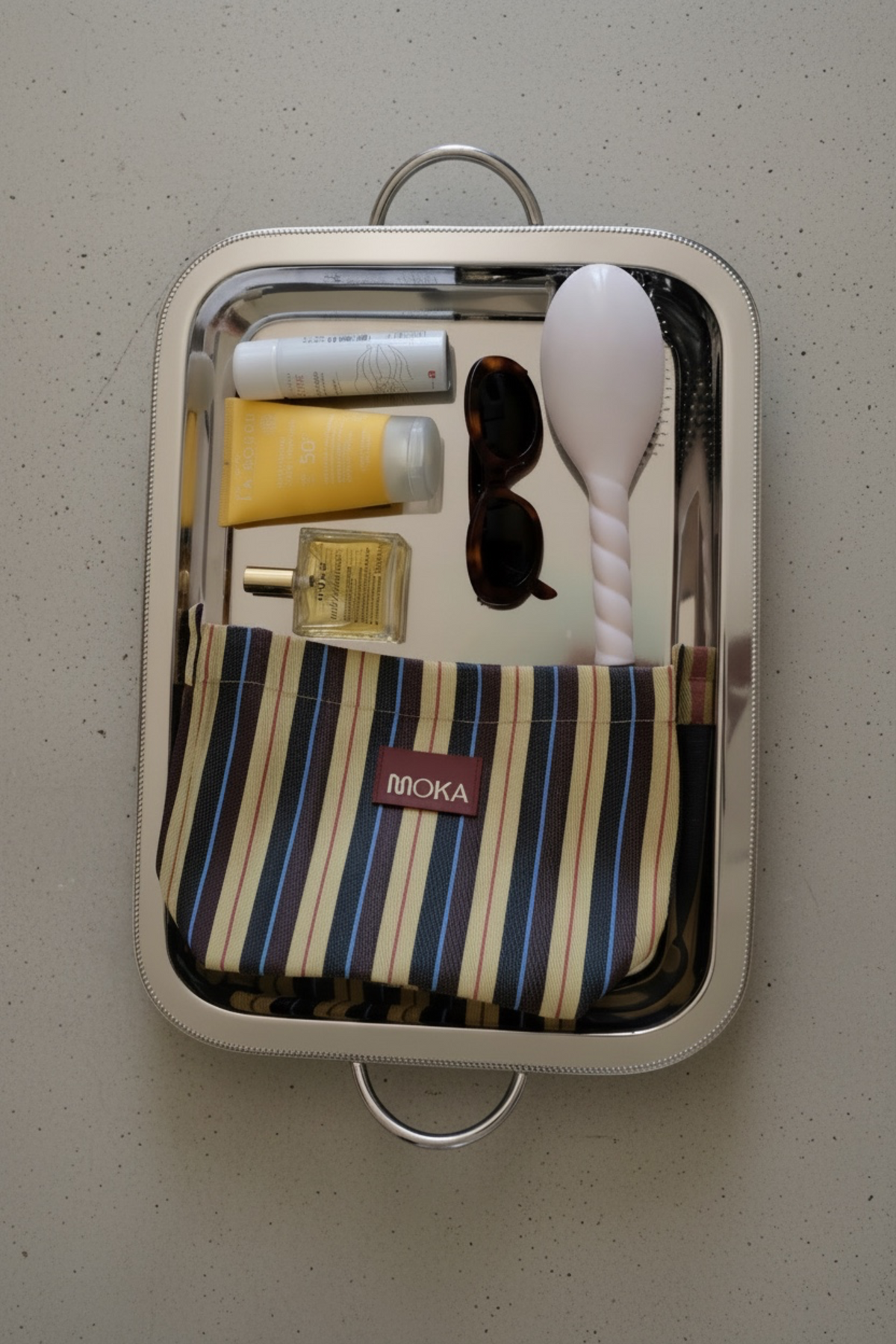 Pochette MOKA Cabanon à rayures colorées posée sur un plateau argenté avec des accessoires de beauté et solaires