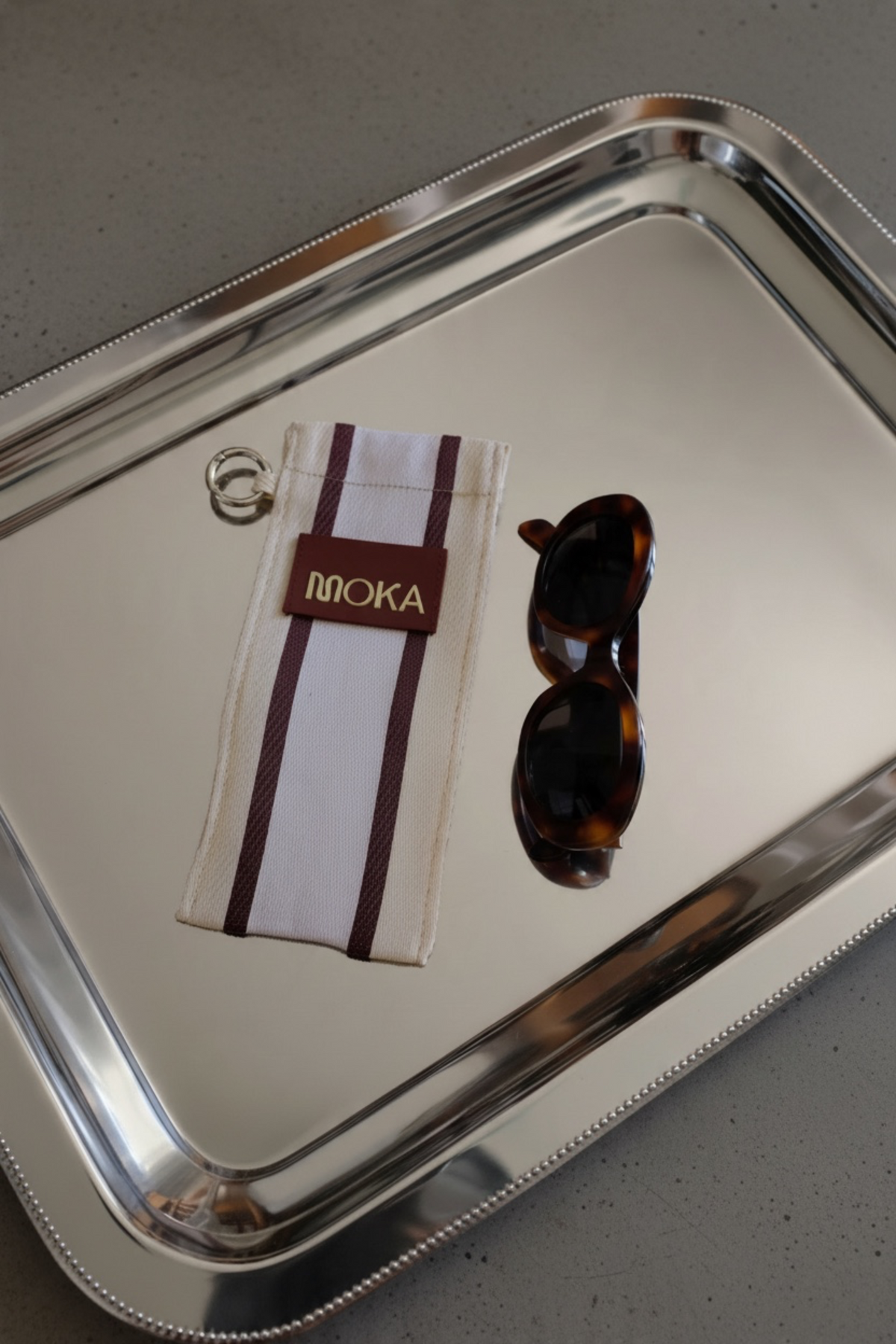 Étui à lunettes MOKA Coconut en coton recyclé rayé beige et bordeaux avec étiquette logo sur un plateau argenté